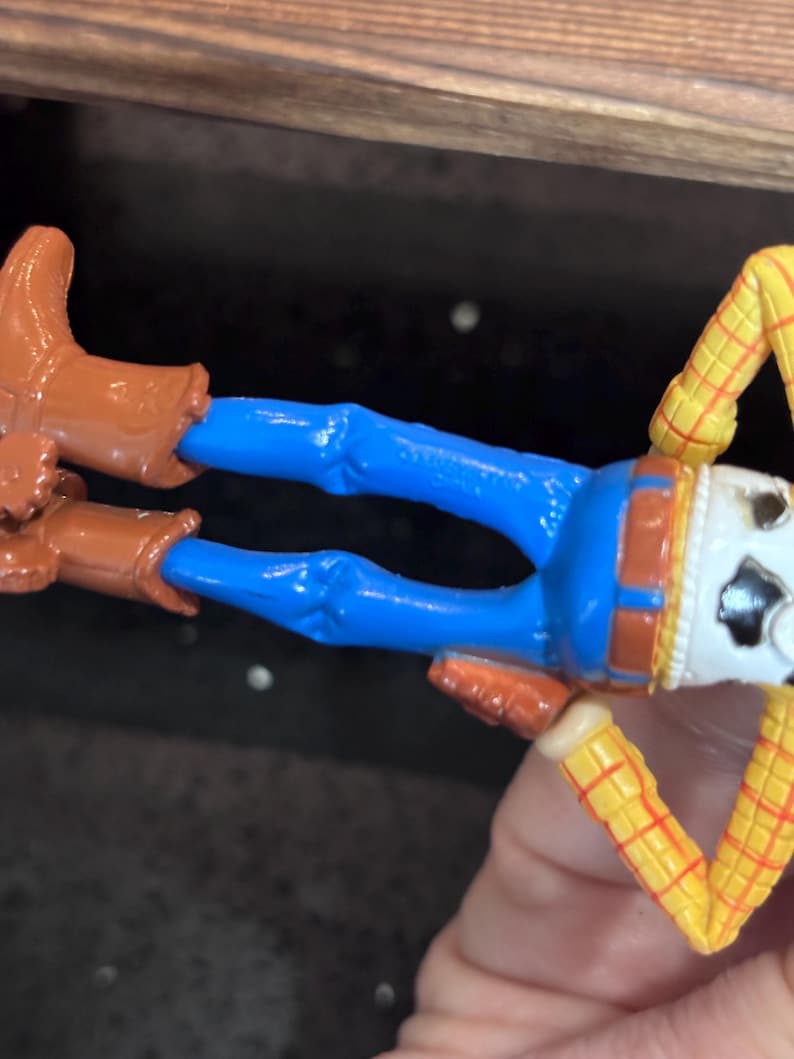 K&ouml;nnte beinhalten: Woody-Actionfigur aus dem Toy Story-Franchise. Die Figur hat eine blaue Hose, braune Stiefel, ein gelb-rotes kariertes Hemd und ein wei&szlig;es Gesicht mit Cowboyhut.