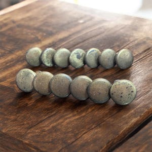 Peut inclure: Deux rangées de boutons de meuble en laiton antique avec une finition mouchetée sont disposées sur une surface en bois foncé. Chaque bouton est rond et a un aspect légèrement vieilli, suggérant une esthétique vintage ou rustique. Les boutons sont espacés uniformément.