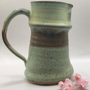 May include: A handmade ceramic pitcher with a light green glaze and brown accents. The pitcher has a wide mouth and a handle. It is sitting on a white surface with pink flowers.