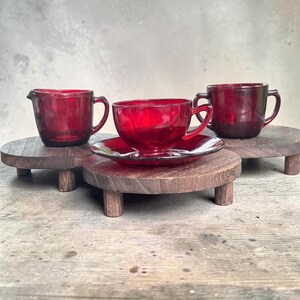 Könnte beinhalten: Ein Set aus drei rubinroten Glasteilen, bestehend aus einem Milchkännchen mit Ausguss, einer Teetasse auf passender Untertasse und einer Zuckerdose mit zwei Henkeln. Jedes Teil besteht aus durchscheinendem rotem Glas auf einem kleinen Holzständer.