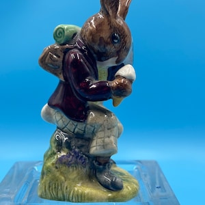May include: A ceramic figurine of a brown rabbit wearing a burgundy jacket and plaid pants, carrying a backpack and holding a mushroom. The rabbit is perched on a grassy base with purple flowers, set against a blue background.