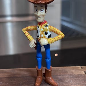 K&ouml;nnte beinhalten: Woody-Actionfigur mit braunem Cowboyhut, gelbem kariertem Hemd, wei&szlig;er Weste mit Sheriff-Abzeichen, blauer Hose und braunen Stiefeln. Die Figur hat einen l&auml;chelnden Gesichtsausdruck und posiert mit den H&auml;nden in den H&uuml;ften.