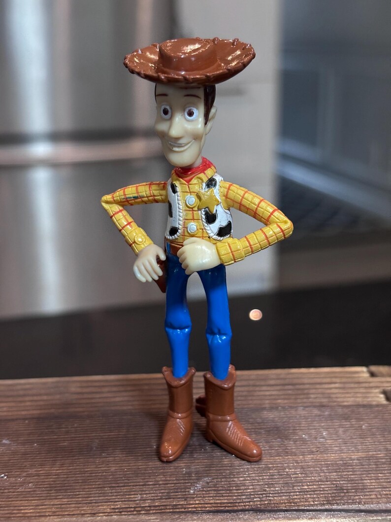 K&ouml;nnte beinhalten: Woody-Actionfigur mit braunem Cowboyhut, gelbem kariertem Hemd, wei&szlig;er Weste mit Sheriff-Abzeichen, blauer Hose und braunen Stiefeln. Die Figur hat einen l&auml;chelnden Gesichtsausdruck und posiert mit den H&auml;nden in den H&uuml;ften.