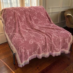 May include: A dusty rose throw blanket featuring a repeating cream-coloured floral pattern across the surface. The rectangular textile is finished with a decorative cream-coloured fringe along the edges, providing a textured border to the fabric.