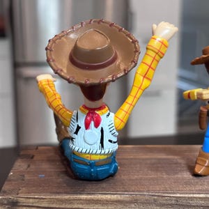 K&ouml;nnte beinhalten: Woody-Actionfigur mit braunem Cowboyhut, gelben karierten &Auml;rmeln und einer wei&szlig;en Weste mit schwarzen Details. Die Figur ist mit erhobenen Armen posiert und zeigt das ikonische Outfit der Figur.