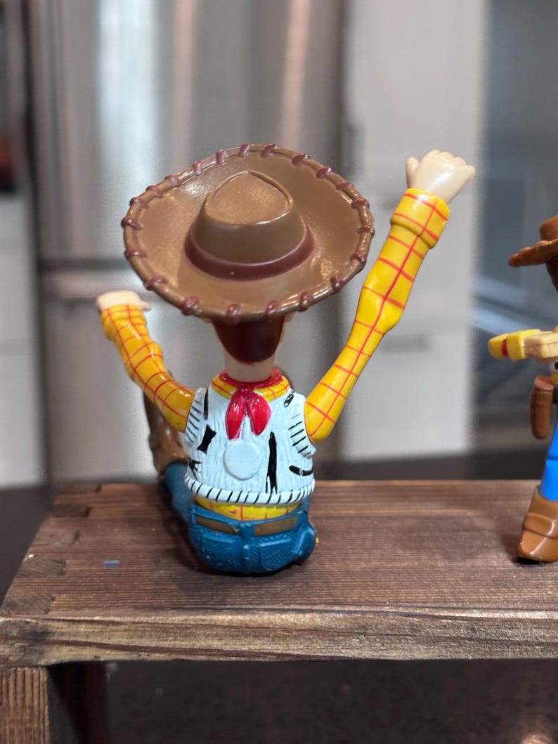 K&ouml;nnte beinhalten: Woody-Actionfigur mit braunem Cowboyhut, gelben karierten &Auml;rmeln und einer wei&szlig;en Weste mit schwarzen Details. Die Figur ist mit erhobenen Armen posiert und zeigt das ikonische Outfit der Figur.