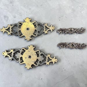 May include: Four antique brass hardware pieces. Two larger pieces feature a central square and ornate filigree edges. Two smaller, matching pieces are also included. The hardware has a tarnished, aged appearance.