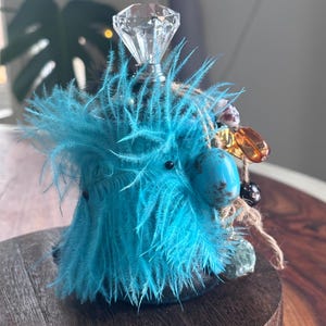 May include: A decorative bottle with a diamond-shaped crystal stopper. The bottle is decorated with bright turquoise feathers, a turquoise stone, and various beads. The bottle sits on a dark wooden stand, creating a unique and artistic display.