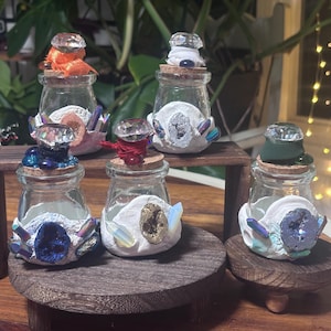 Op de afbeelding: Een verzameling decoratieve glazen potten met kristallen kurkproppen. Elke pot is versierd met kleurrijke kristallen en geode-achtige accenten. De potten worden tentoongesteld op kleine houten platforms, wat een mystiek of apotheekthema suggereert.