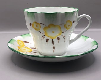 May include: A white teacup and saucer with a green and yellow floral design. The cup has a scalloped rim and a handle. The saucer has a green stripe around the edge.