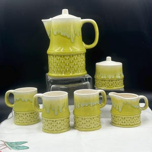 Puede incluir: Un juego de café de cerámica vintage en tonos verdes y blancos. El juego incluye una cafetera, azucarero, lechera y cuatro tazas. Las piezas tienen un diseño de tejido de cesta texturizado en la parte inferior y un efecto de esmalte goteante.