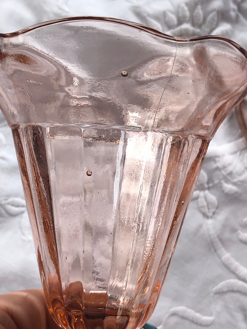 May include: A pink glass vase with a fluted design and a scalloped rim. The vase is made of clear glass with a pink tint.