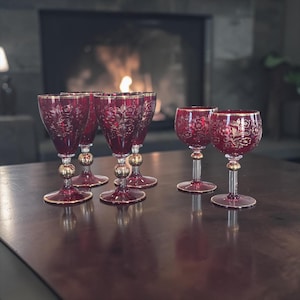Puede incluir: Juego de seis copas de vino rojo rubí con diseños florales dorados. Cada copa tiene un borde dorado y un detalle de bola dorada en el tallo. Las copas están dispuestas sobre una mesa marrón oscuro.
