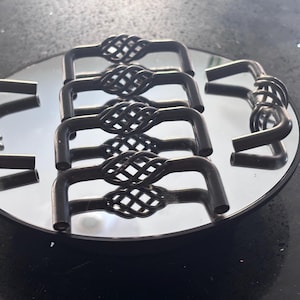 Puede incluir: Un conjunto de seis tiradores de armario de metal oscuro con un diseño decorativo tejido en el centro. Los tiradores están dispuestos sobre una superficie espejada, reflejando su forma y el entorno.