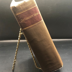 Op de afbeelding: Een bruin boek met de titel "Old Curiosity Shop" op de rug. Het boek staat op een gouden metalen standaard.