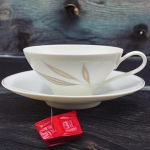 Peut inclure: Une tasse à thé et une soucoupe blanches avec un motif de feuilles dorées. La tasse à thé a une anse et la soucoupe a un bord doré. Un sachet de thé rouge est suspendu à la soucoupe.