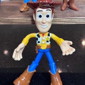 K&ouml;nnte beinhalten: Woody-Actionfigur mit braunem Cowboyhut, gelbem Hemd, blauer Hose und braunen Stiefeln. Die Figur hat ein Sheriff-Abzeichen und ein Holster. Das Spielzeug ist ein Sammlerst&uuml;ck aus der Toy Story-Reihe.