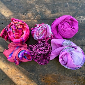 May include: Six rolled scarves in various shades of pink and purple. The collection includes a bright pink plaid scarf, a multi-colored pastel scarf, a solid magenta scarf, a coral and blue floral scarf, a black and pink leopard print scarf, and a lavender floral scarf.