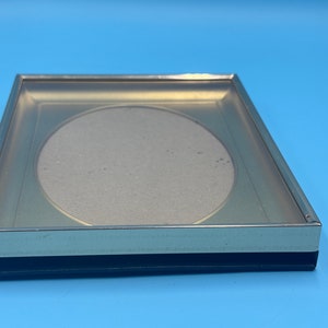 Puede incluir: Un marco de fotos plateado con un borde dorado y una abertura circular para una foto. El marco está sobre una superficie azul.