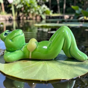 May include: A glossy green ceramic frog figurine lying on its back with hands behind its head. The frog has a smooth, reflective surface and a light yellow bikini top detail. It is positioned horizontally, showcasing a relaxed, reclining posture.