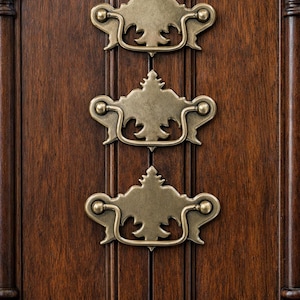 Peut inclure: Gros plan sur une porte d'armoire en bois avec trois poignées de tiroir en laiton antique. Les poignées ont un design orné et sont montées verticalement sur le bois brun foncé.