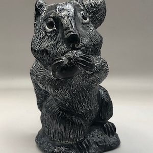 Puede incluir: Una talla de ardilla en piedra de jabón negra que sostiene una nuez en sus patas. La ardilla está sentada sobre una base y tiene una apariencia detallada y realista.