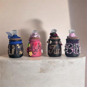 May include: Four decorative jars with crystal lids. The jars are painted in black, pink, and blue, and decorated with flowers, ribbons, and text. The jars are displayed on a white surface. The jars are likely for decorative purposes.