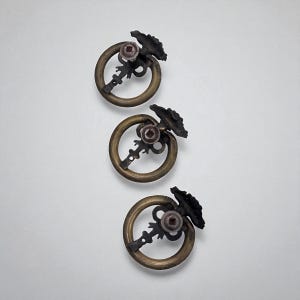 Op de afbeelding: Drie antieke messing ringtrekkers met sierlijke, donkere metalen achterplaten. Elke trekker heeft een cirkelvormige ring, een decoratief midden en een gestileerd sleutelvormig element.