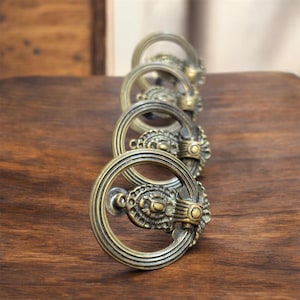 以下が含まれることがあります： 円形ハンドルデザインのアンティーク真鍮製引き出しプル。各プルは、花柄の装飾的で詳細なフェースプレートを備えています。木製の表面に配置され、ヴィンテージ感を演出しています。