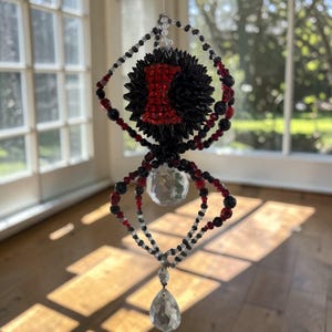 Puede incluir: Adorno decorativo de araña con cuerpo negro y detalles rojos. La araña presenta una bola de cristal y un colgante de cristal en forma de lágrima. Las patas están hechas de cuentas negras, rojas y blancas. La araña cuelga frente a una ventana.