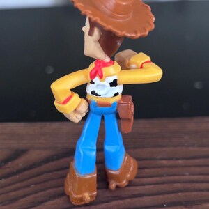 K&ouml;nnte beinhalten: Kleine Woody-Actionfigur aus Kunststoff. Die Figur tr&auml;gt einen braunen Cowboyhut, ein gelbes Hemd, eine blaue Jeans und braune Stiefel. Ein rotes Halstuch und ein Holster sind ebenfalls zu sehen.
