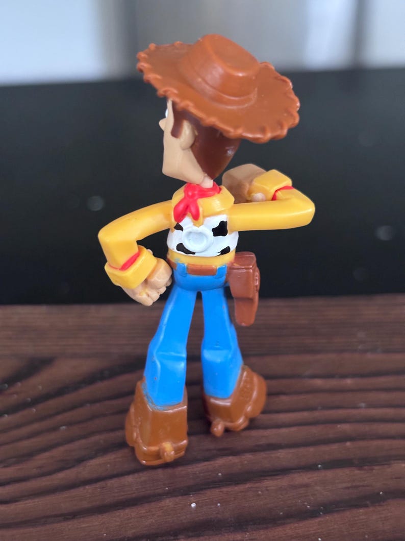 K&ouml;nnte beinhalten: Kleine Woody-Actionfigur aus Kunststoff. Die Figur tr&auml;gt einen braunen Cowboyhut, ein gelbes Hemd, eine blaue Jeans und braune Stiefel. Ein rotes Halstuch und ein Holster sind ebenfalls zu sehen.