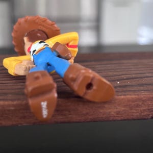 K&ouml;nnte beinhalten: Eine Spielzeugfigur einer Figur aus dem Animationsfilm Toy Story, die auf einer Holzoberfl&auml;che liegt. Die Figur tr&auml;gt einen braunen Cowboyhut, ein gelbes Hemd, blaue Hosen und braune Stiefel. Die Figur befindet sich in einer entspannten Pose.