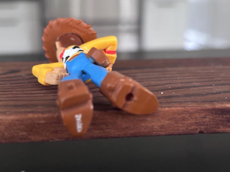 K&ouml;nnte beinhalten: Eine Spielzeugfigur einer Figur aus dem Animationsfilm Toy Story, die auf einer Holzoberfl&auml;che liegt. Die Figur tr&auml;gt einen braunen Cowboyhut, ein gelbes Hemd, blaue Hosen und braune Stiefel. Die Figur befindet sich in einer entspannten Pose.