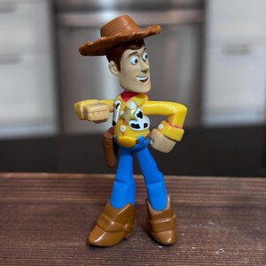 K&ouml;nnte beinhalten: Eine Woody-Actionfigur aus dem Toy Story-Franchise. Die Figur tr&auml;gt einen braunen Cowboyhut, ein gelbes Hemd, eine blaue Hose und braune Stiefel. Die Figur hat ein rotes Halstuch und ein Holster mit einer Spielzeugpistole.