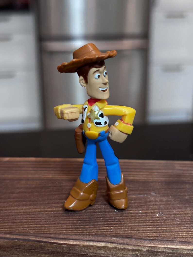 K&ouml;nnte beinhalten: Eine Woody-Actionfigur aus dem Toy Story-Franchise. Die Figur tr&auml;gt einen braunen Cowboyhut, ein gelbes Hemd, eine blaue Hose und braune Stiefel. Die Figur hat ein rotes Halstuch und ein Holster mit einer Spielzeugpistole.