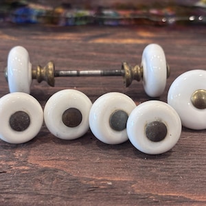 Peut inclure: Ensemble de boutons de meubles en céramique blanche avec des détails en laiton. Les boutons ronds ont un centre sombre et sont disposés sur une surface en bois foncé. Un ensemble comprend une tige de connexion.