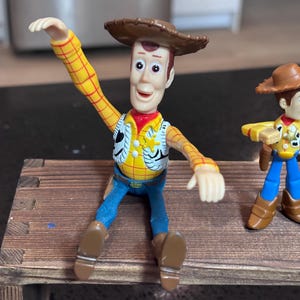 K&ouml;nnte beinhalten: Woody-Actionfigur aus dem Toy Story-Franchise. Die Figur tr&auml;gt ein gelbes kariertes Hemd, eine blaue Jeans, ein rotes Halstuch und einen braunen Cowboyhut. Die Figur sitzt auf einer kleinen Holzbank.