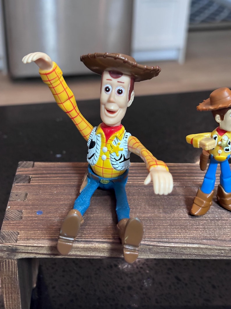 K&ouml;nnte beinhalten: Woody-Actionfigur aus dem Toy Story-Franchise. Die Figur tr&auml;gt ein gelbes kariertes Hemd, eine blaue Jeans, ein rotes Halstuch und einen braunen Cowboyhut. Die Figur sitzt auf einer kleinen Holzbank.