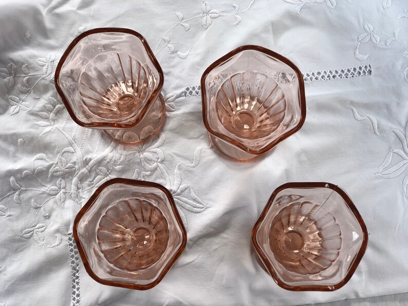 May include: Four pink glass dessert cups with a fluted design. The cups are arranged in a square pattern on a white tablecloth.