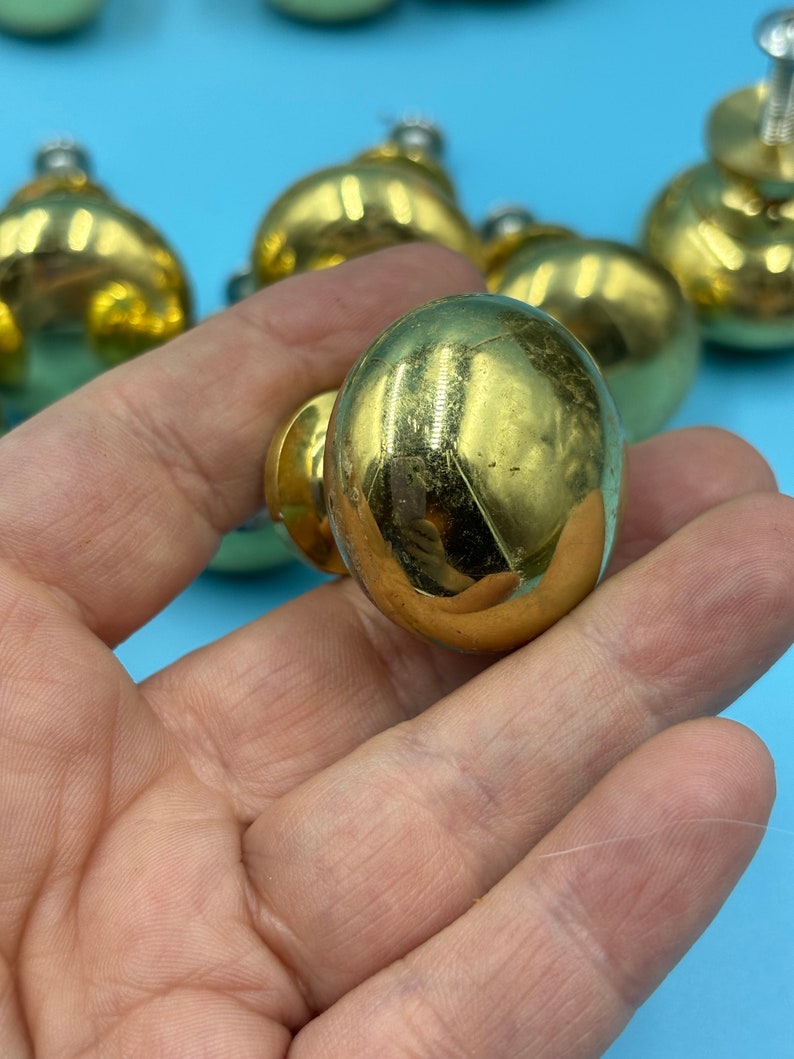 May include: A close-up of a gold-toned cabinet knob with a round, slightly domed shape. The knob is smooth and shiny, with a slight sheen.