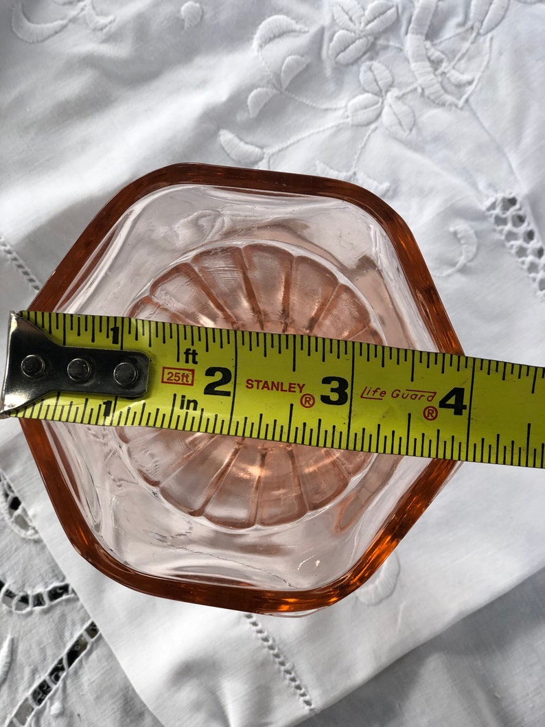 May include: A pink glass dish with a scalloped edge, measuring approximately 3 inches in diameter. The dish is sitting on a white fabric surface.