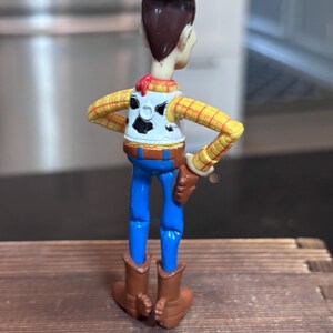 K&ouml;nnte beinhalten: Woody-Actionfigur, von hinten gesehen, mit braunem Cowboyhut, gelbem kariertem Hemd, blauer Jeans und braunen Stiefeln. Die Figur hat eine wei&szlig;e Weste mit schwarzem Kuhfellmuster und einem roten Halstuch. Die Figur steht auf einer Holzoberfl&auml;che.