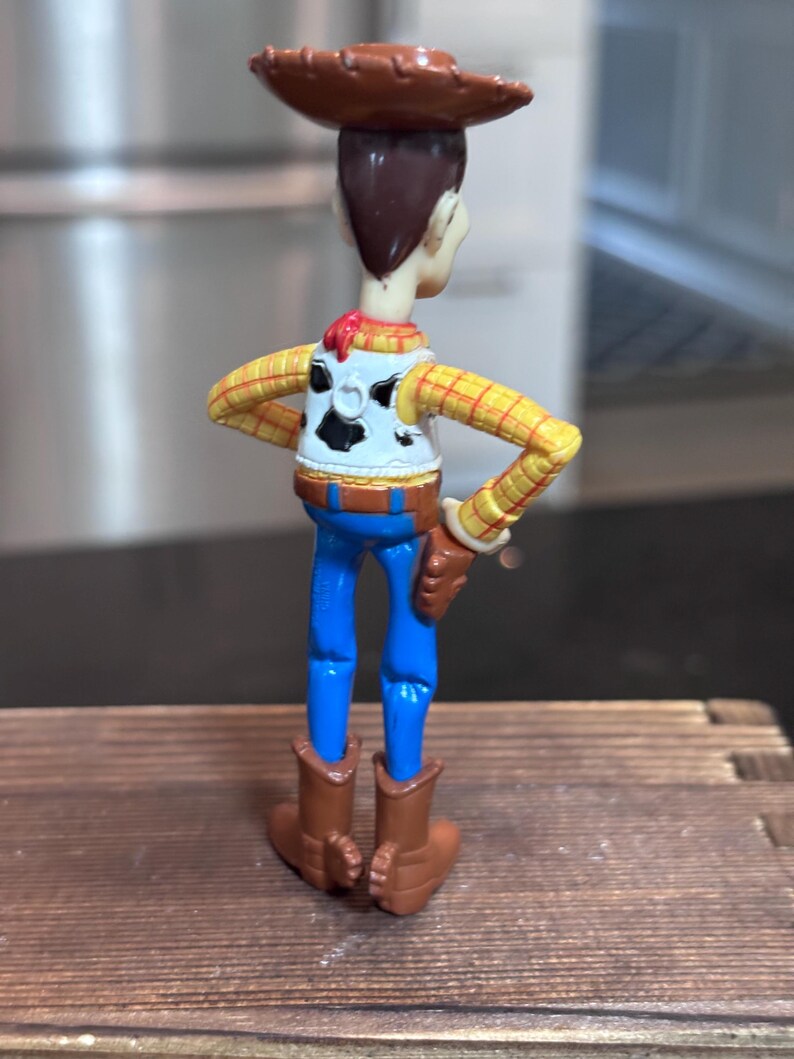 K&ouml;nnte beinhalten: Woody-Actionfigur, von hinten gesehen, mit braunem Cowboyhut, gelbem kariertem Hemd, blauer Jeans und braunen Stiefeln. Die Figur hat eine wei&szlig;e Weste mit schwarzem Kuhfellmuster und einem roten Halstuch. Die Figur steht auf einer Holzoberfl&auml;che.