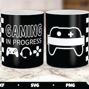 May include: A black and white mug with the text "Gaming in Progress" and a video game controller, headset, and directional pad. The mug is on a white surface.