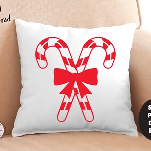 K&ouml;nnte beinhalten: Ein wei&szlig;es Kissen mit einem roten und wei&szlig;en Zuckerstangenmuster. Die Zuckerstangen sind gekreuzt und mit einer roten Schleife zusammengebunden. Das Kissen liegt auf einem beigen Sofa.