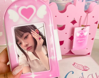 May include: A pink plastic photo frame with a heart and wings design. The frame has a photo of a young woman with dark hair. The frame is sitting on a white surface with a pink and white bag in the background.