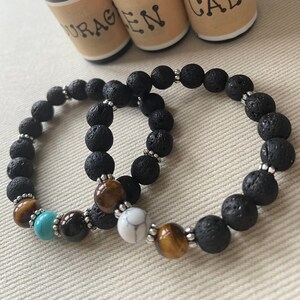May include: Two black lava stone bracelets with tiger eye, turquoise, and white beads. The bracelets are next to three small glass bottles with labels that read "Courage", "Zen", and "Calm".