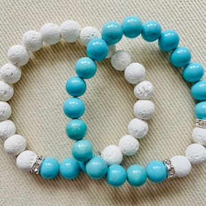 May include: Two bracelets, one with white lava rock beads and the other with turquoise beads. Both bracelets have a silver charm with rhinestones.