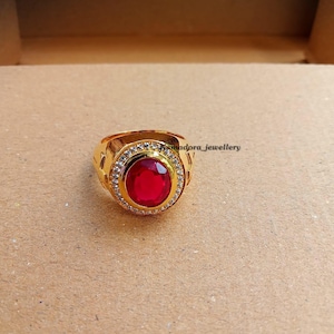 Könnte beinhalten: Ein goldfarbener Ring mit einem großen, ovalen, roten Edelstein in der Mitte. Der Edelstein ist von einem Kranz aus kleineren, klaren Steinen umgeben. Der Ring hat ein strukturiertes Design. Der Text "smdora_jewellery" ist sichtbar.